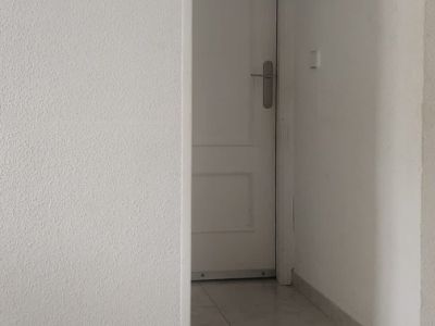 Zicht op een lichte hal met lichtgekleurde tegelvloer en witte gestructureerde muren. Een decoratief cherubijnornament siert de linkermuur, leidend naar een witte deur met zilveren handgreep.