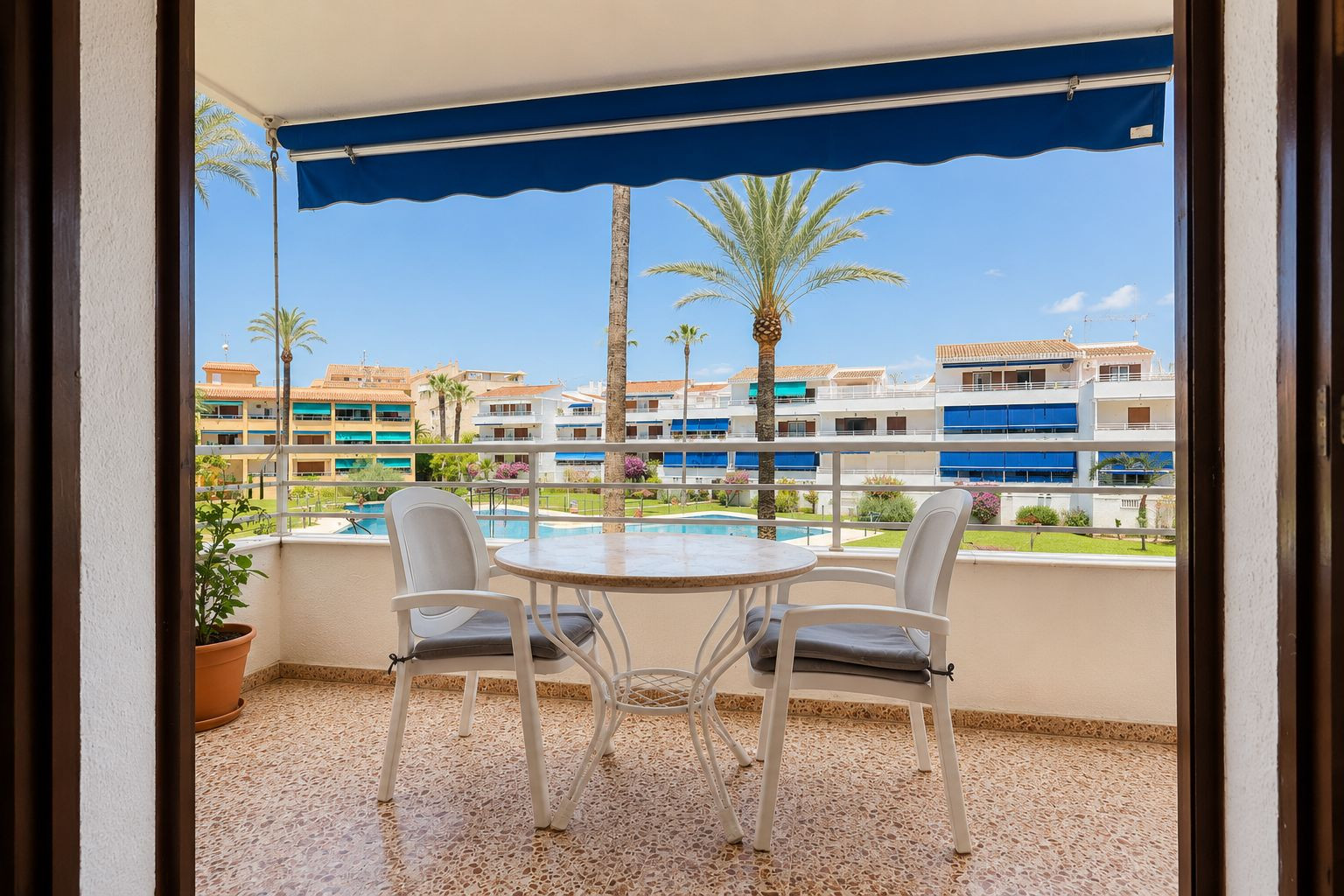 Betegeld terras met een ronde tafel en twee stoelen, onder een blauw zonnescherm. Biedt uitzicht op het gemeenschappelijke zwembad en palmbomen.