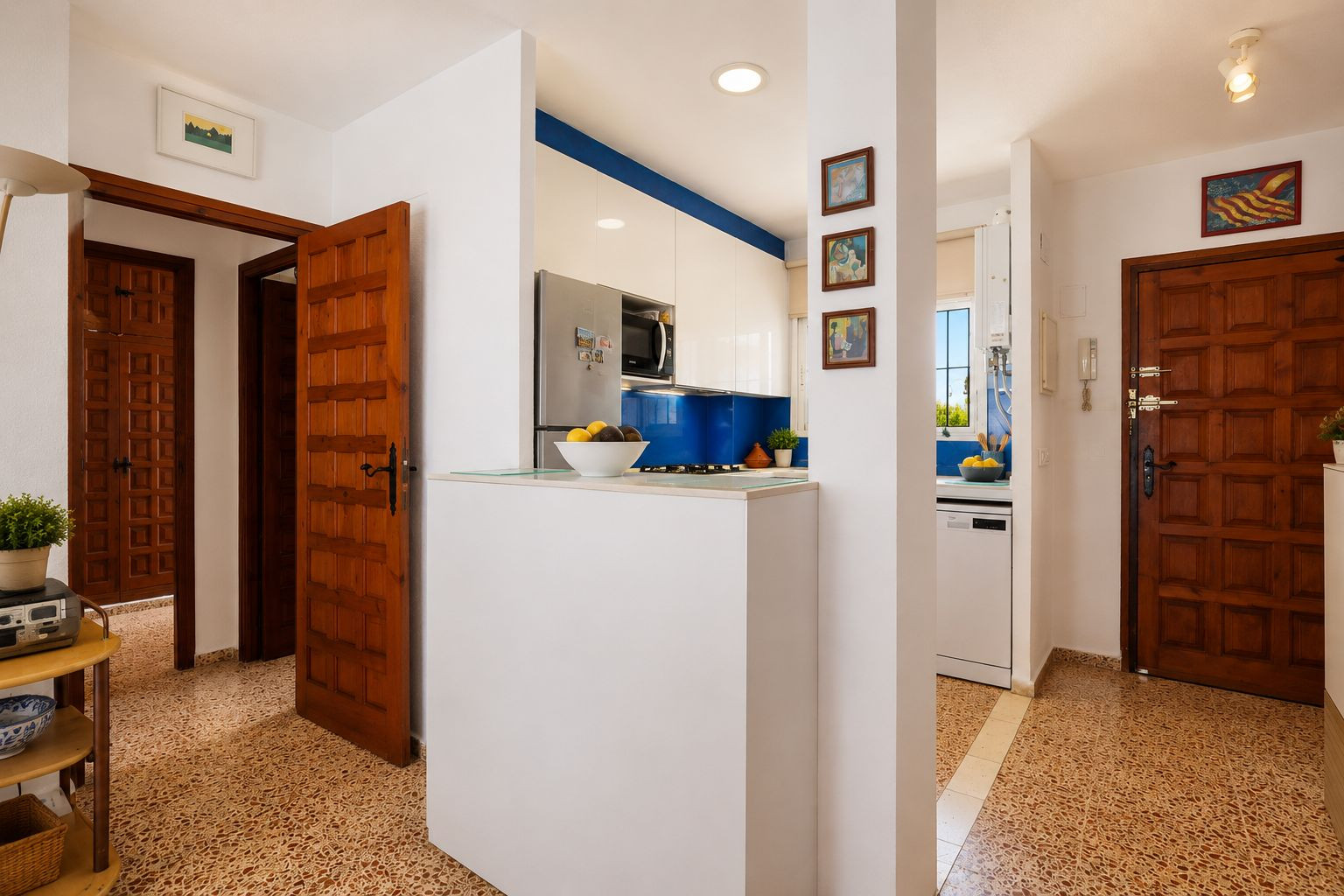 De entreehal toont opvallende vloertegels met patroon en klassieke houten deuren. Een moderne keuken met blauwe achterwand en witte kasten is zichtbaar, gescheiden door een functionele bar.