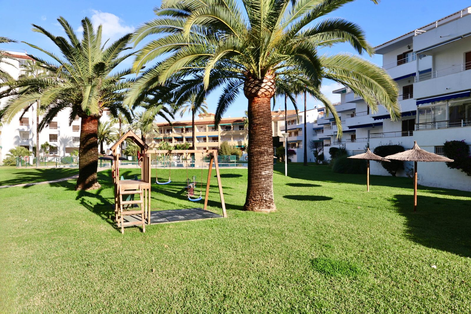 Grote gemeenschappelijke tuin met groen gazon, houten speeltuin, volwassen palmbomen en twee strooien parasols, omringd door appartementen.