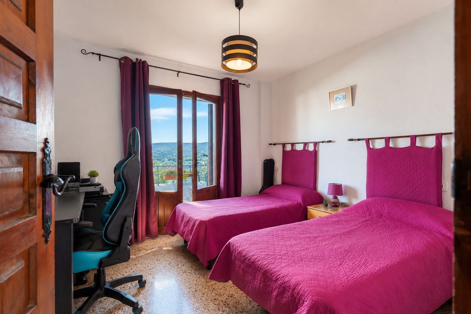 Lichte slaapkamer met twee eenpersoonsbedden, fuchsia hoofdeinden en directe toegang tot een balkon met berguitzicht. Voorzien van een bureau en terrazzovloer.