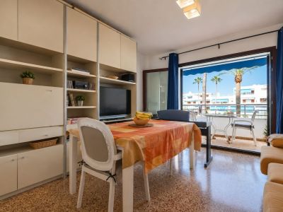 Lichte woon-eetkamer met eettafel en ingebouwde kast, direct grenzend aan een terras met uitzicht op palmbomen en witte gebouwen.