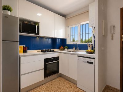 Moderne en functionele keuken met glanzend witte kasten, een licht werkblad en een blauwe achterwand. Voorzien van inbouwapparatuur en een raam met uitzicht op groen.
