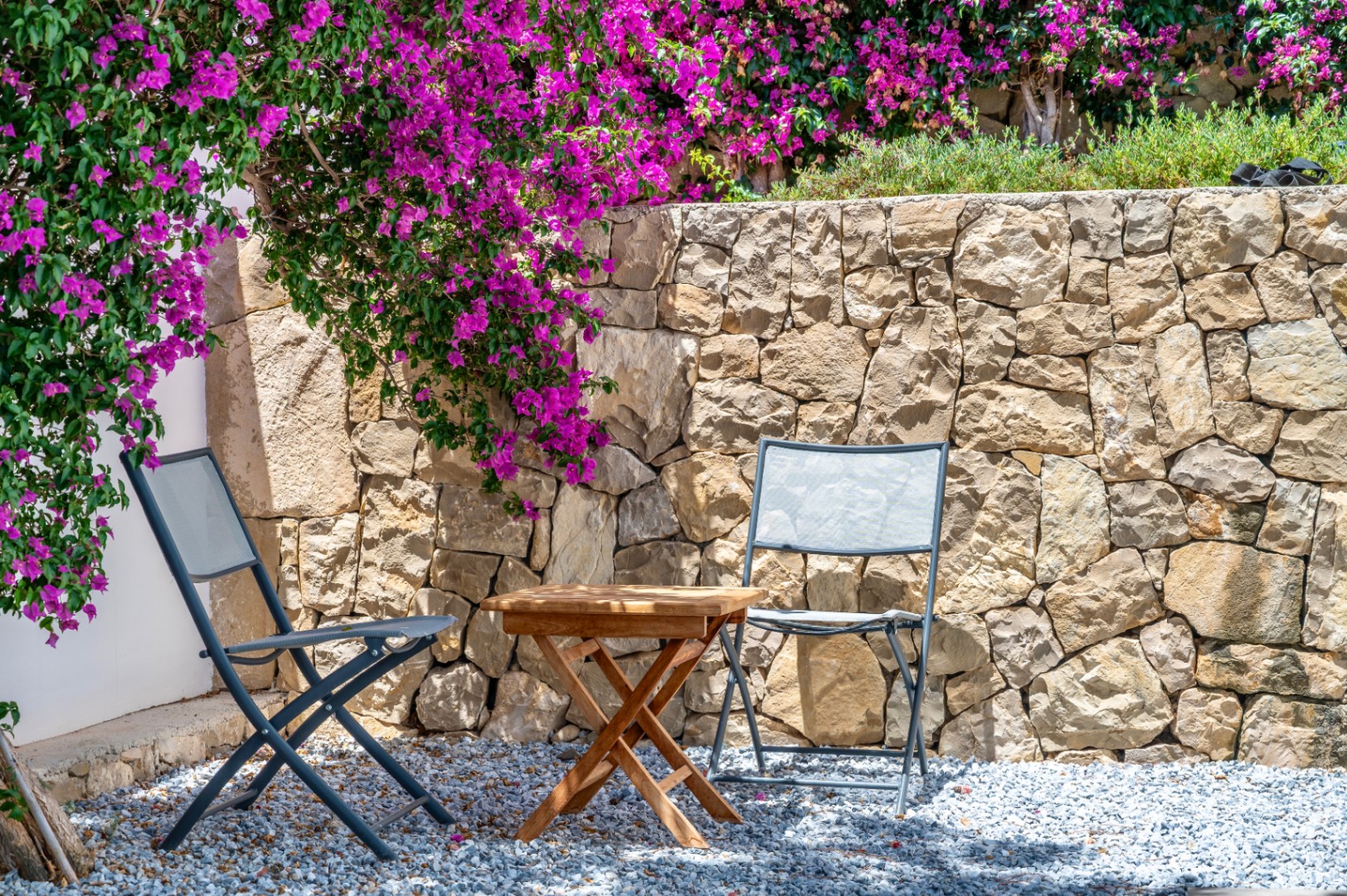 Buiten zithoek met twee grijze klapstoelen en een houten tafel op grind. Een natuurstenen muur is versierd met levendige magenta bougainvillea. Buiten zithoek met twee grijze klapstoelen en een houten tafel op grind. Een natuurstenen muur is versierd met levendige magenta bougainvillea.