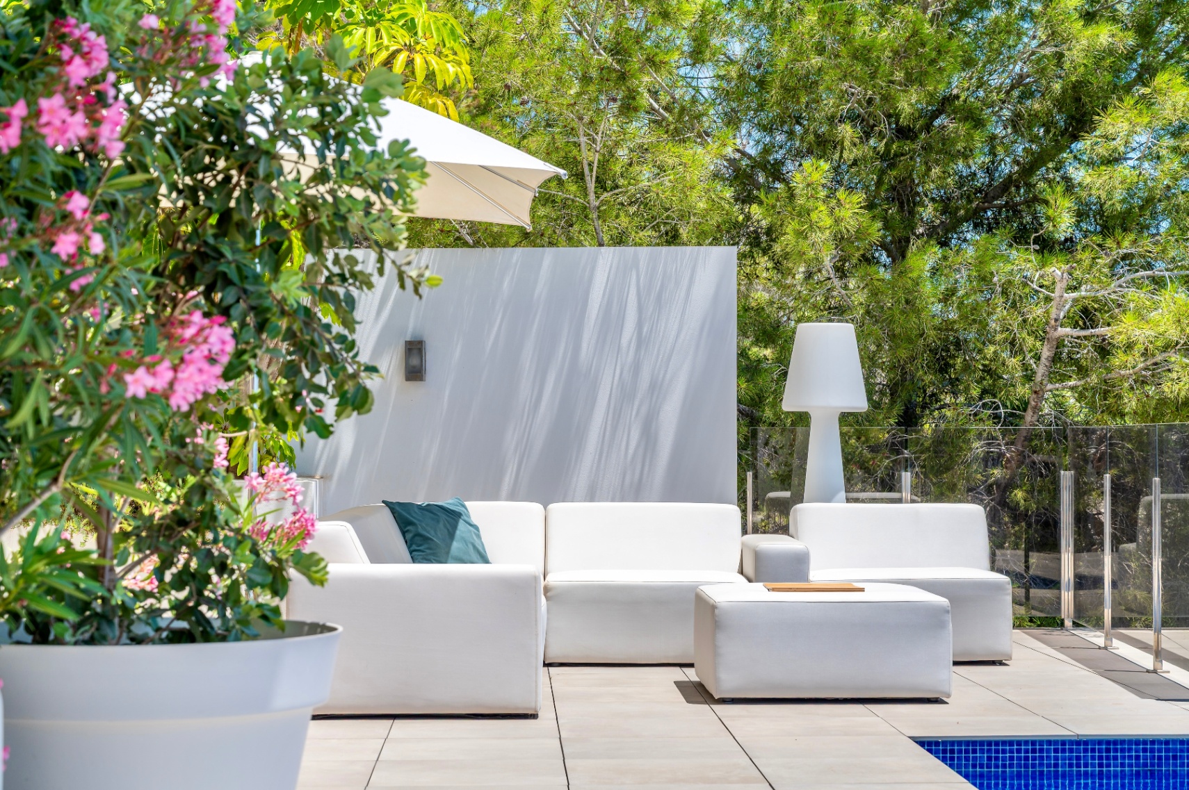 Hedendaagse buitenlounge met witte modulaire banken en bijpassende poef op een betegeld terras. Omgeven door weelderig groen, een glazen balustrade en een roze bloeiende plant. Hedendaagse buitenlounge met witte modulaire banken en bijpassende poef op een betegeld terras. Omgeven door weelderig groen, een glazen balustrade en een roze bloeiende plant.