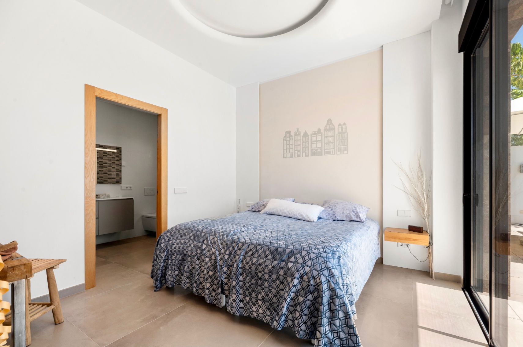 Lichte slaapkamer met tweepersoonsbed en een accentmuur met gebouwensilhouetten. Directe toegang tot een en-suite badkamer en een terras via een grote glazen schuifdeur. Lichte slaapkamer met tweepersoonsbed en een accentmuur met gebouwensilhouetten. Directe toegang tot een en-suite badkamer en een terras via een grote glazen schuifdeur.