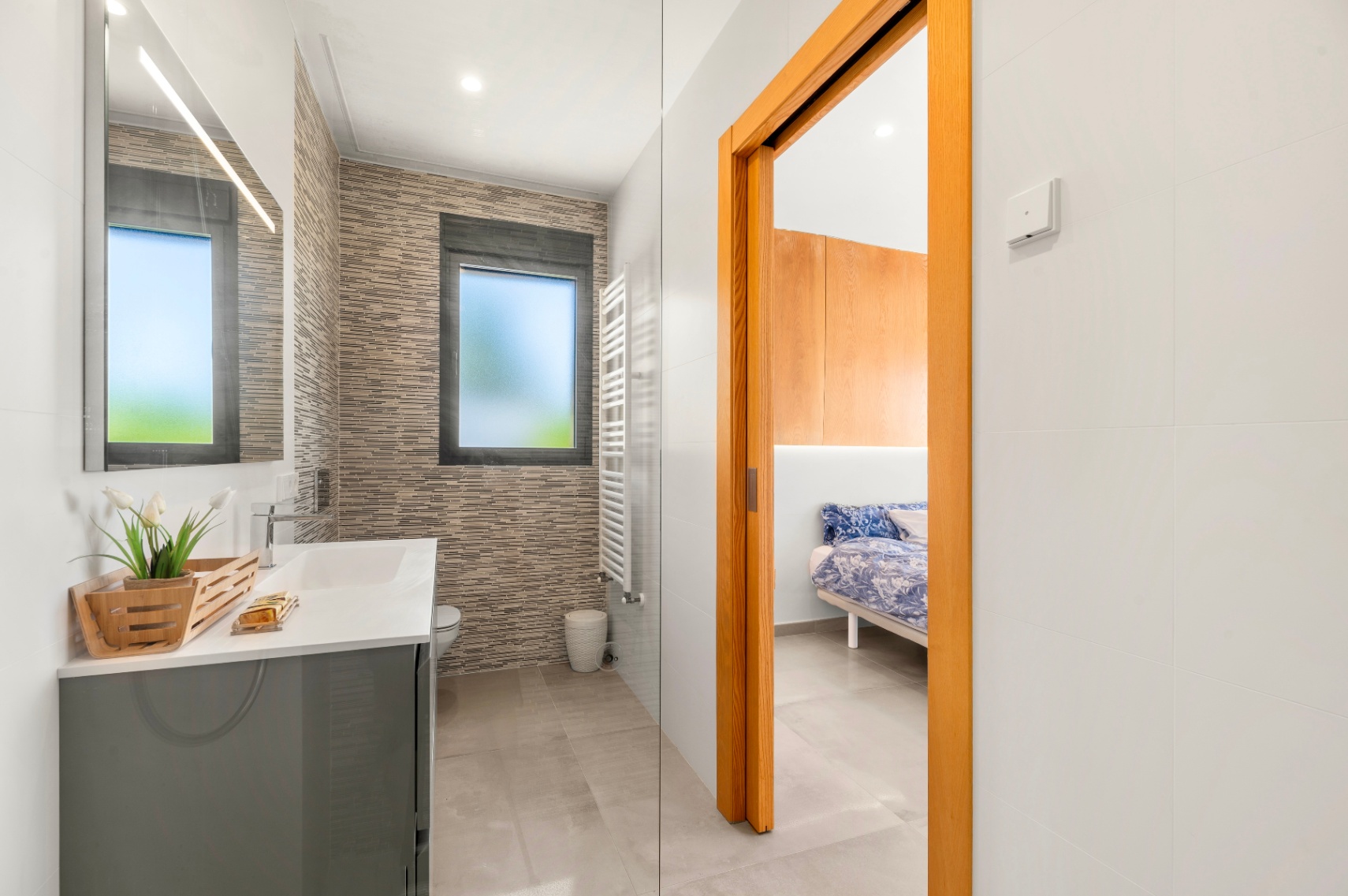 Moderne badkamer met grijze wastafel, verlichte spiegel en getextureerde wandtegels. Een matglazen raam en een witte handdoekradiator zijn zichtbaar, grenzend aan een slaapkamer. Moderne badkamer met grijze wastafel, verlichte spiegel en getextureerde wandtegels. Een matglazen raam en een witte handdoekradiator zijn zichtbaar, grenzend aan een slaapkamer.