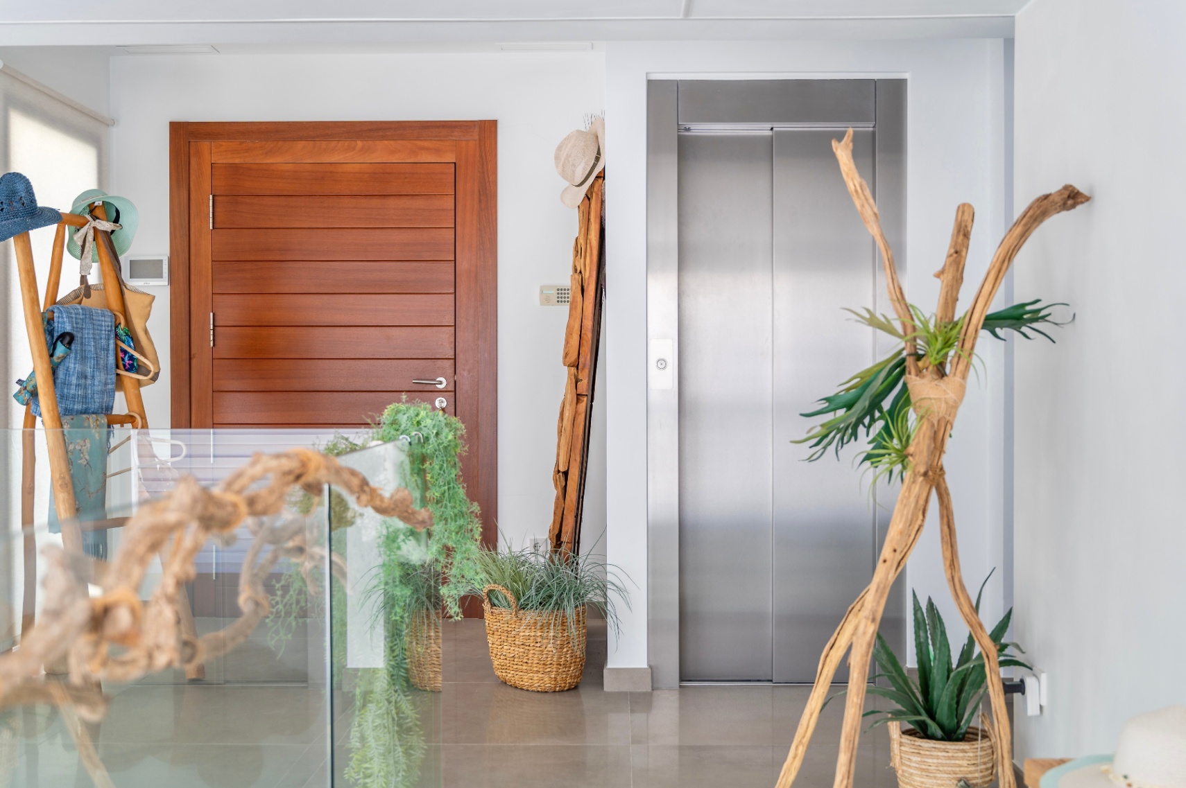 Moderne binnenhal met een strakke houten deur en een roestvrijstalen lift. De ruimte is elegant gedecoreerd met natuurlijke elementen zoals drijfhout, planten in manden en een glazen balustrade. Moderne binnenhal met een strakke houten deur en een roestvrijstalen lift. De ruimte is elegant gedecoreerd met natuurlijke elementen zoals drijfhout, planten in manden en een glazen balustrade.