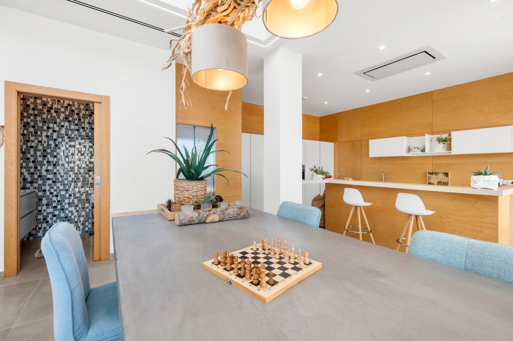 Moderne eetruimte met grijze tafel en lichtblauwe stoelen. Een unieke hanglamp van natuurlijk hout en een open keuken met lichte houten panelen zijn zichtbaar. Toegang tot een mozaïekte badkamer. Moderne eetruimte met grijze tafel en lichtblauwe stoelen. Een unieke hanglamp van natuurlijk hout en een open keuken met lichte houten panelen zijn zichtbaar. Toegang tot een mozaïekte badkamer.