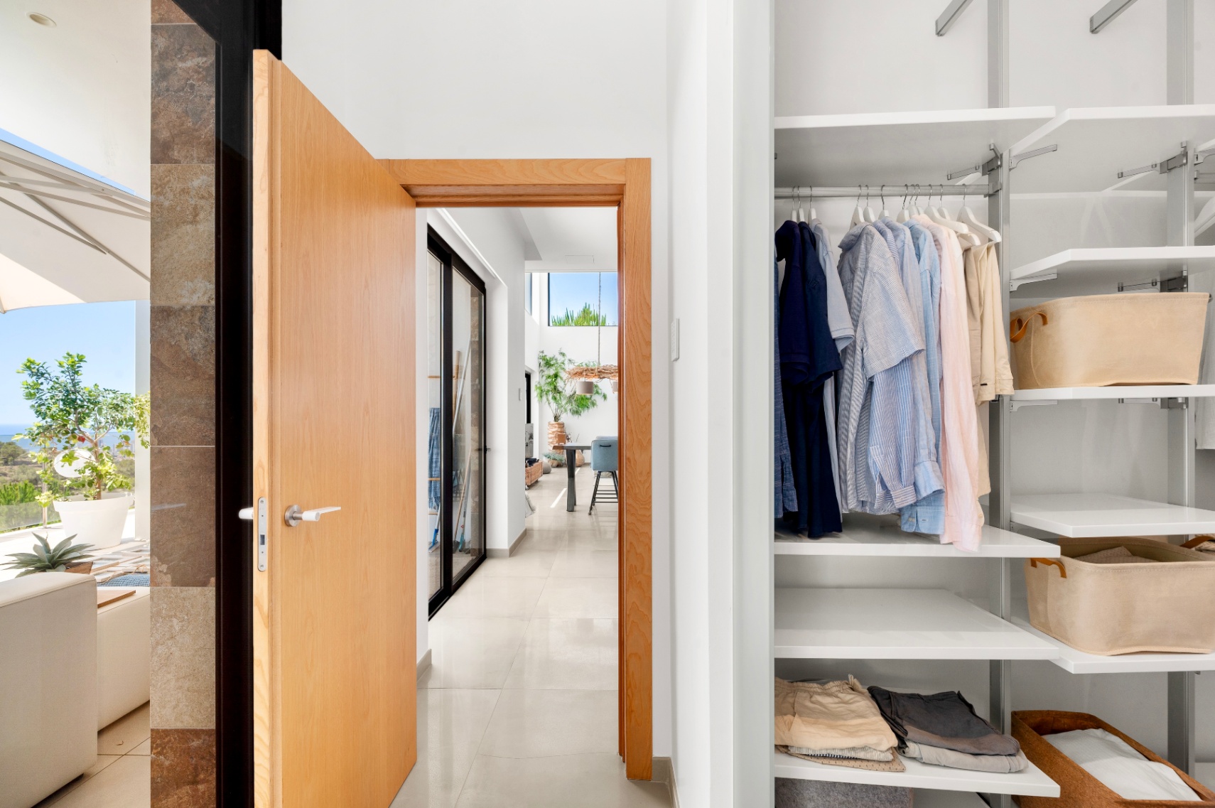 Moderne halingang met open lichte houten deur, die een ingebouwde witte kledingkast met verstelbare planken en een zonnig terras met zeezicht onthult. Moderne halingang met open lichte houten deur, die een ingebouwde witte kledingkast met verstelbare planken en een zonnig terras met zeezicht onthult.