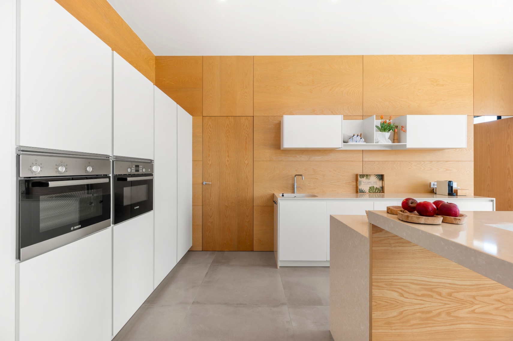 Moderne keuken met geïntegreerde witte kasten en Bosch-ovens. Het centrale eiland met licht stenen werkblad en de lichte houten wand- en plafondbekleding kenmerken de ruimte. Moderne keuken met geïntegreerde witte kasten en Bosch-ovens. Het centrale eiland met licht stenen werkblad en de lichte houten wand- en plafondbekleding kenmerken de ruimte.