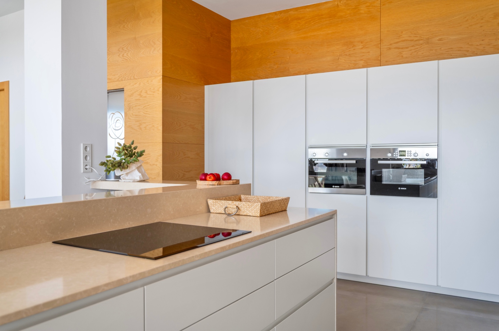 Moderne keuken met zandkleurig werkblad en geïntegreerde inductiekookplaat. Witte greeploze kasten herbergen twee ingebouwde Bosch-ovens en een houten accentmuur. Moderne keuken met zandkleurig werkblad en geïntegreerde inductiekookplaat. Witte greeploze kasten herbergen twee ingebouwde Bosch-ovens en een houten accentmuur.