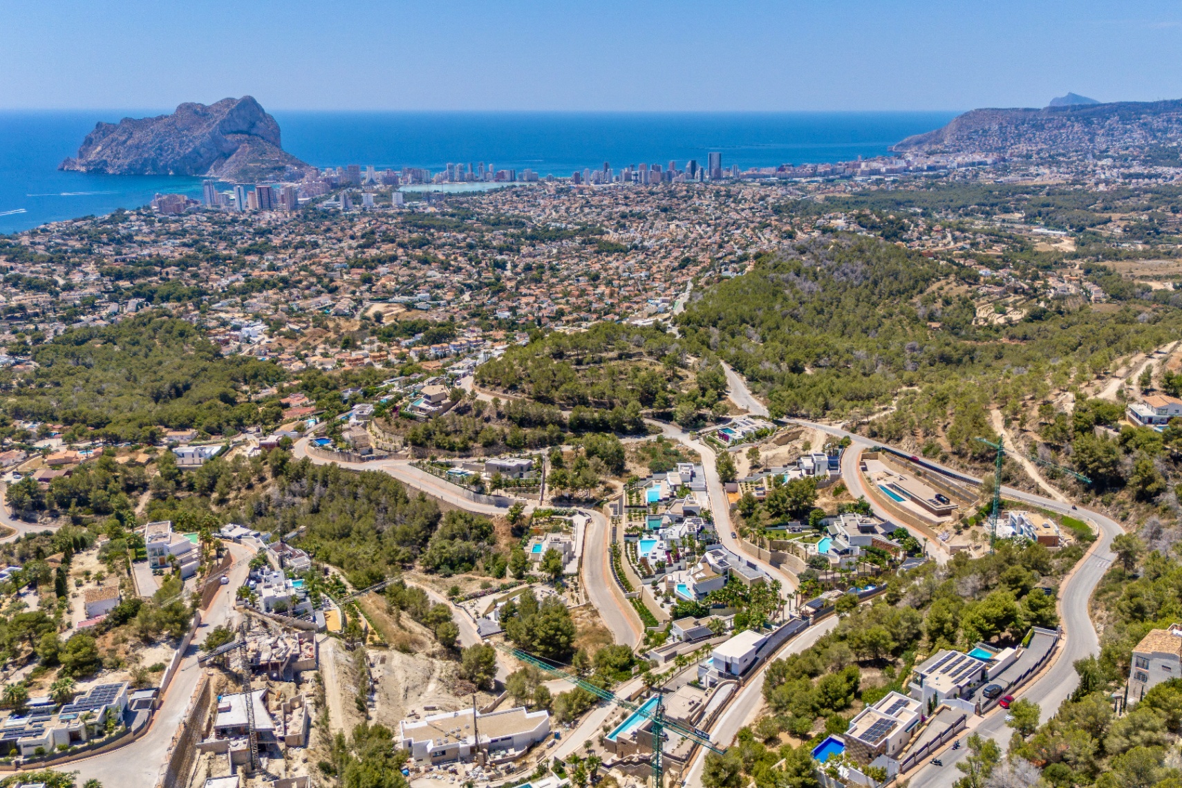 Panoramisch luchtbeeld van Calpe, met de Peñon de Ifach, de kustlijn en een residentiële wijk met villa's en zwembaden tussen groene heuvels en kronkelwegen. Panoramisch luchtbeeld van Calpe, met de Peñon de Ifach, de kustlijn en een residentiële wijk met villa's en zwembaden tussen groene heuvels en kronkelwegen.