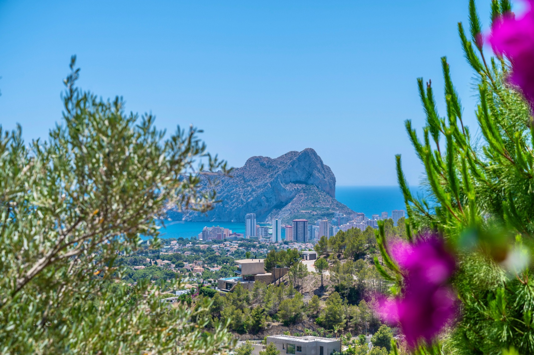 Panoramisch uitzicht op de Peñón de Ifach en de kustlijn van Calpe, omlijst door mediterrane begroeiing, met moderne villa's op de heuvel. Panoramisch uitzicht op de Peñón de Ifach en de kustlijn van Calpe, omlijst door mediterrane begroeiing, met moderne villa's op de heuvel.