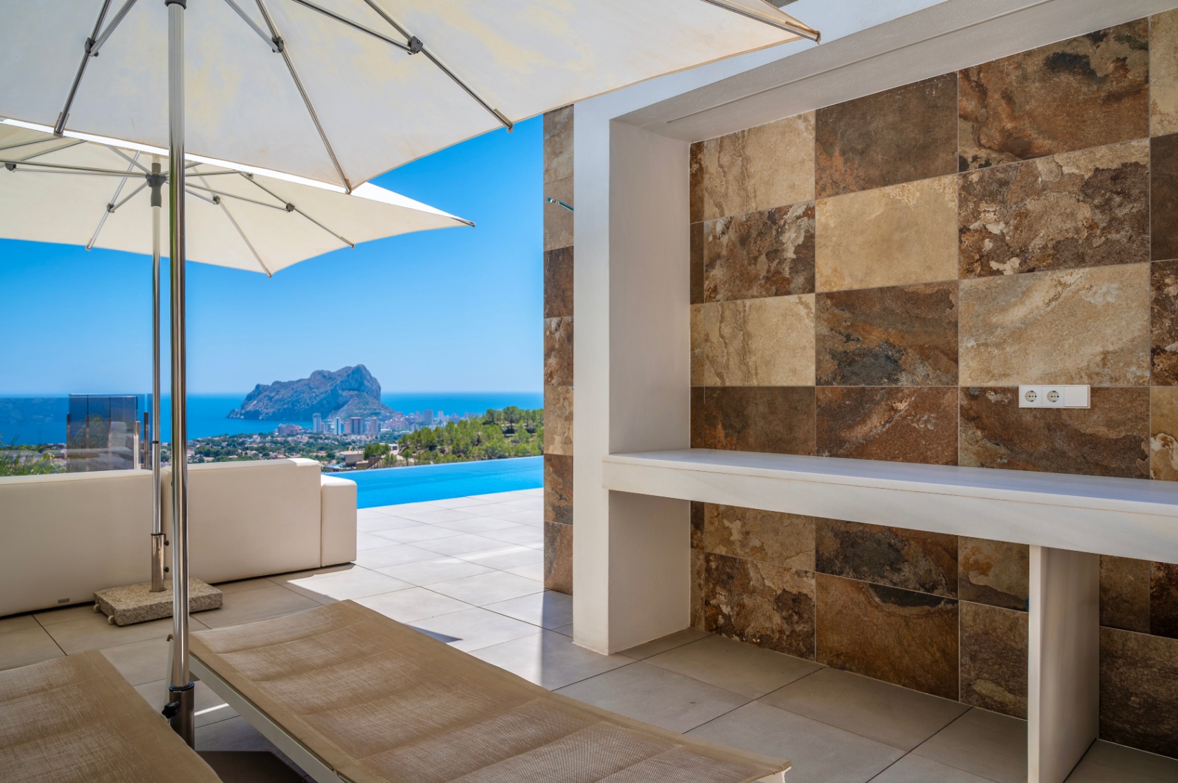 Ruim buitenterras met ingebouwde balie, afgewerkt met natuursteen-look tegels in diverse aardetinten. Panoramisch uitzicht op zee, de infinity pool en de Peñon de Ifach. Ruim buitenterras met ingebouwde balie, afgewerkt met natuursteen-look tegels in diverse aardetinten. Panoramisch uitzicht op zee, de infinity pool en de Peñon de Ifach.