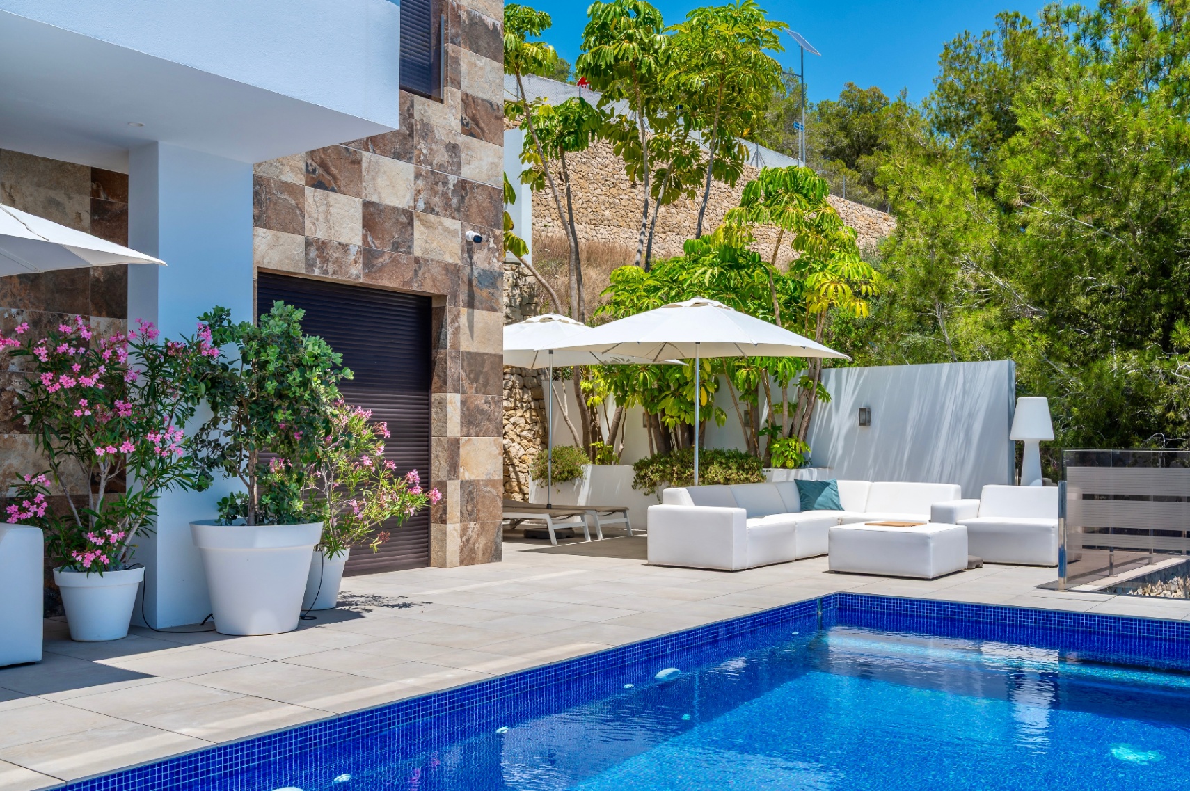 Ruim terras met blauw mozaïekzwembad en wit buitenmeubilair onder parasols, aangevuld met een stenen gevel en weelderige mediterrane beplanting. Ruim terras met blauw mozaïekzwembad en wit buitenmeubilair onder parasols, aangevuld met een stenen gevel en weelderige mediterrane beplanting.