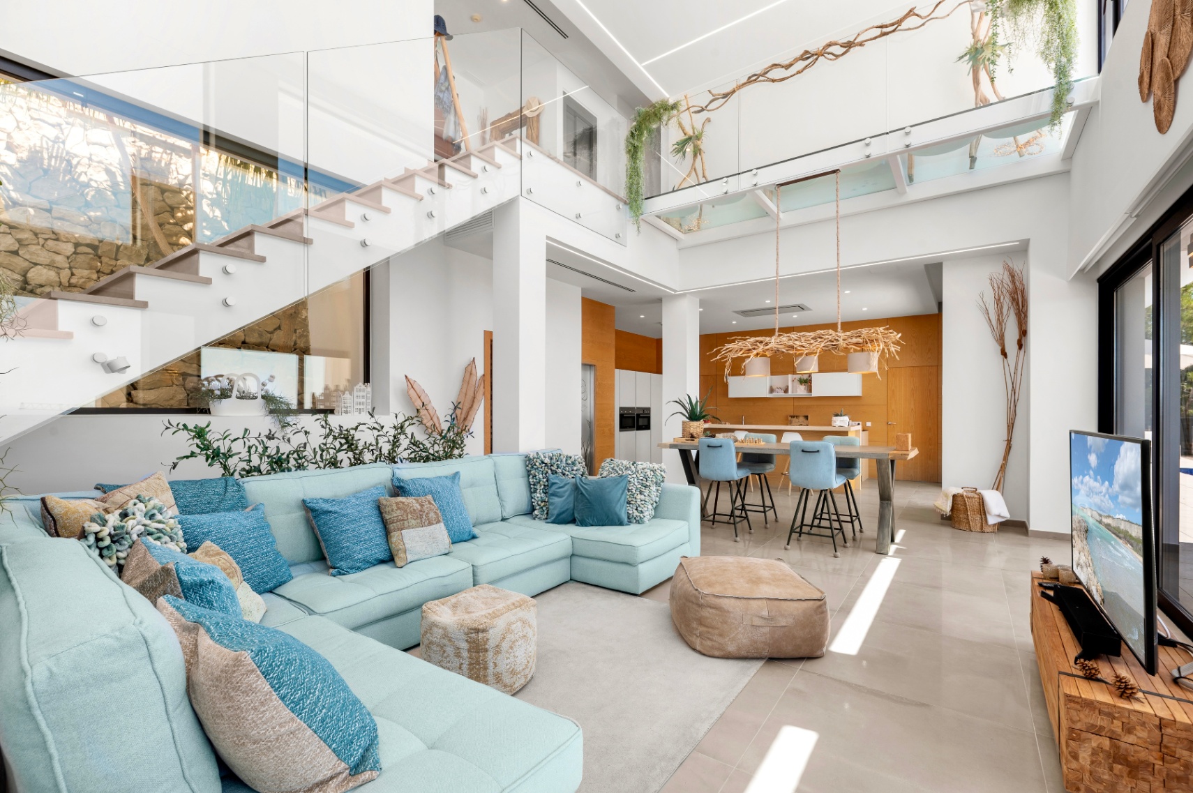 Ruime open woonkamer met lichtblauwe hoekbank, moderne glazen trap en keuken met warme houten accenten, rijkelijk verlicht door natuurlijk lichtinval. Ruime open woonkamer met lichtblauwe hoekbank, moderne glazen trap en keuken met warme houten accenten, rijkelijk verlicht door natuurlijk lichtinval.