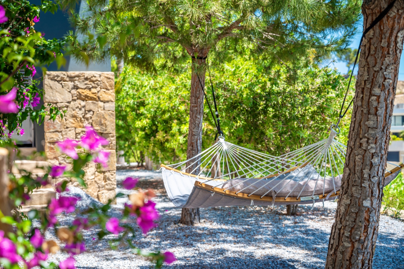 Rustige tuinhoek met een comfortabele grijze hangmat met houten spreidstokken, tussen bomen op een grindbed, omlijst door levendige bougainvillea. Rustige tuinhoek met een comfortabele grijze hangmat met houten spreidstokken, tussen bomen op een grindbed, omlijst door levendige bougainvillea.