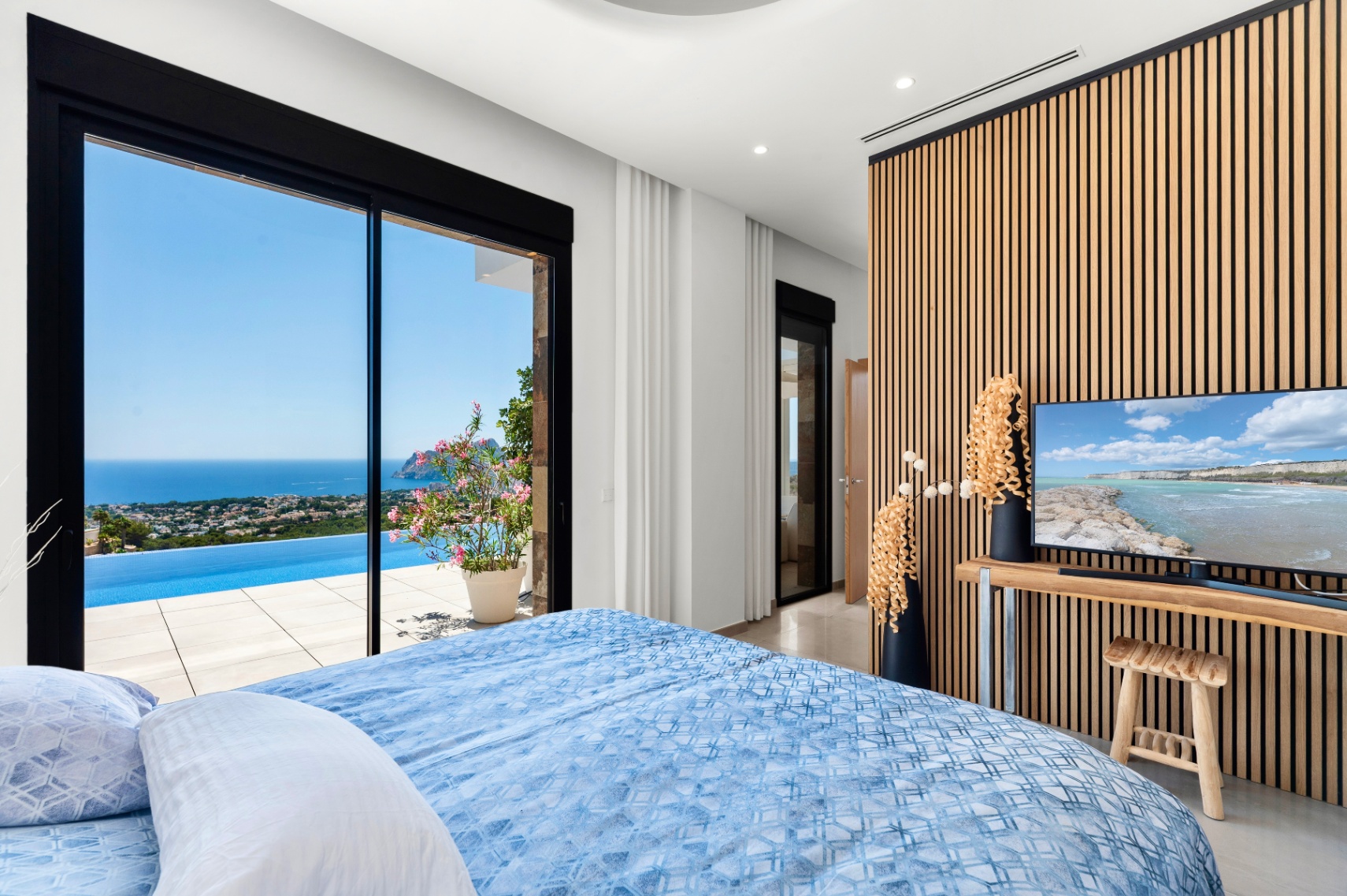 Slaapkamer met grote glazen schuifdeuren naar een infinity pool en panoramisch zeezicht. Een moderne houten lattenwand vormt de achtergrond voor de tv, passend bij het lichte interieur. Slaapkamer met grote glazen schuifdeuren naar een infinity pool en panoramisch zeezicht. Een moderne houten lattenwand vormt de achtergrond voor de tv, passend bij het lichte interieur.