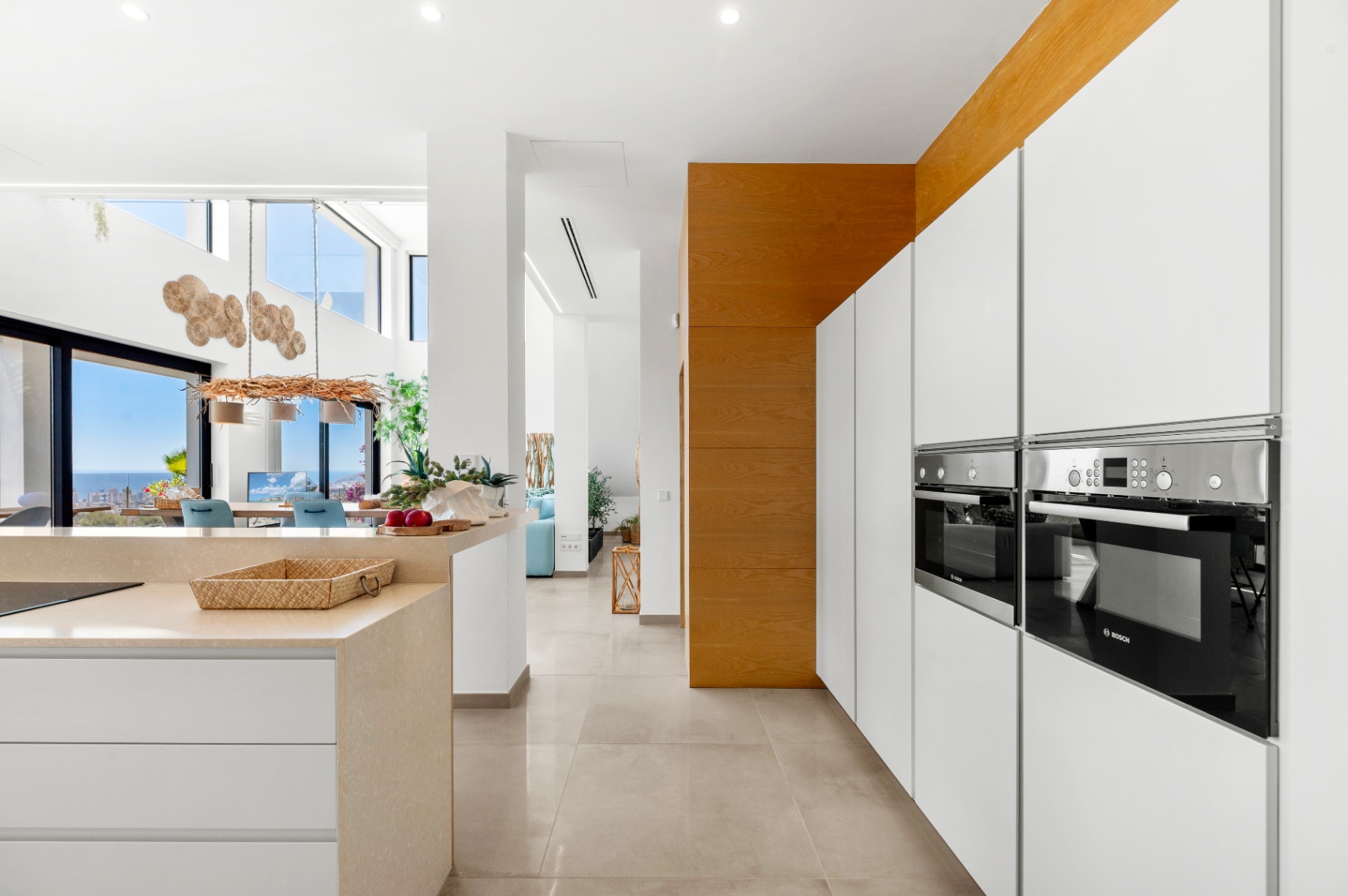 Strakke moderne keuken met geïntegreerde Bosch ovens, witte greeploze kasten en een licht stenen eiland. De open indeling verbindt met een eetruimte met panoramisch zeezicht via glazen deuren. Strakke moderne keuken met geïntegreerde Bosch ovens, witte greeploze kasten en een licht stenen eiland. De open indeling verbindt met een eetruimte met panoramisch zeezicht via glazen deuren.