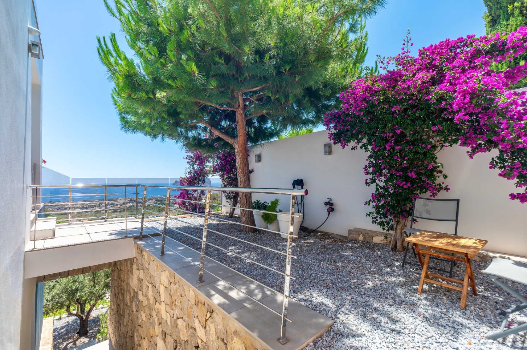 Terras met moderne RVS balustrade en glas, biedt panoramisch zeezicht. Aangrenzende grindtuin met grote dennenboom en levendige paarse bougainvillea tegen een witte muur. Terras met moderne RVS balustrade en glas, biedt panoramisch zeezicht. Aangrenzende grindtuin met grote dennenboom en levendige paarse bougainvillea tegen een witte muur.