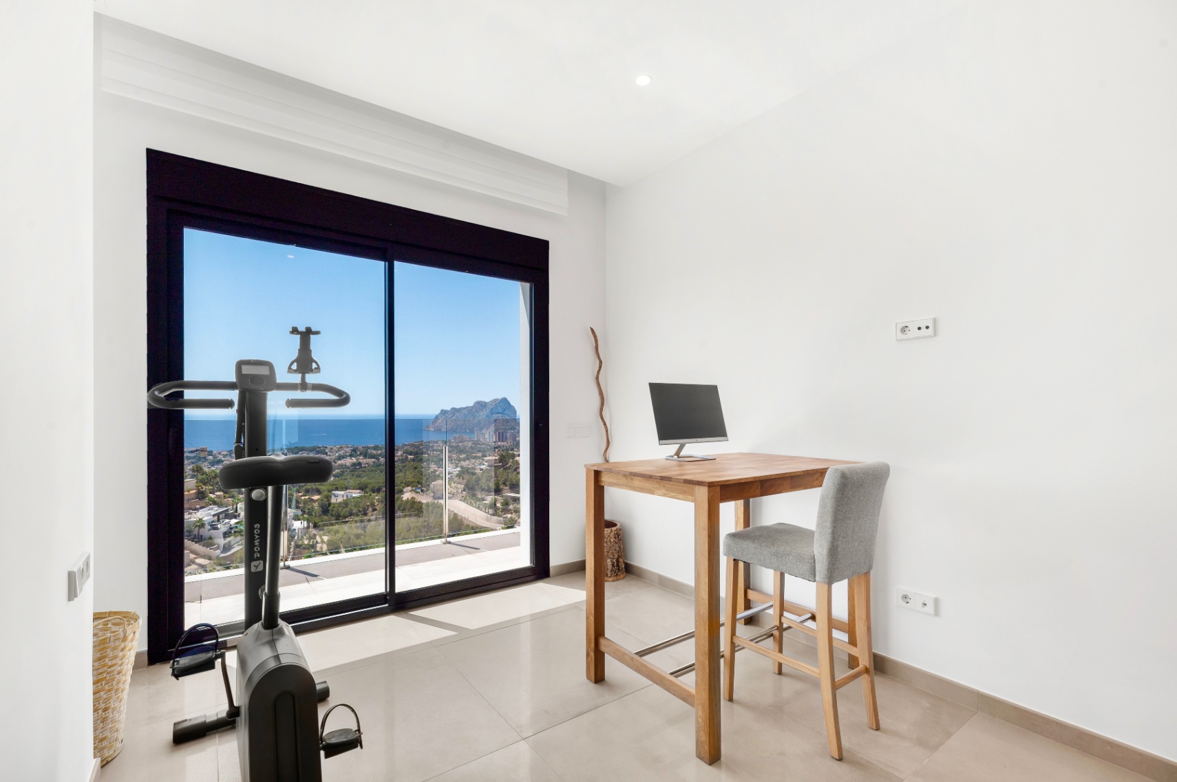 Veelzijdige kamer met houten hoog bureau en hometrainer, gericht op het weidse uitzicht op zee en de Peñon de Ifach door de grote glazen schuifdeuren. Veelzijdige kamer met houten hoog bureau en hometrainer, gericht op het weidse uitzicht op zee en de Peñon de Ifach door de grote glazen schuifdeuren.