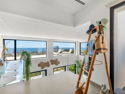 Bovenverdieping met lichte tegels en glazen balustrade. Grote ramen bieden panoramisch uitzicht op zee en de Peñon de Ifach. Kenmerken omvatten een glazen loopbrug met ovale treden en een houten rek.