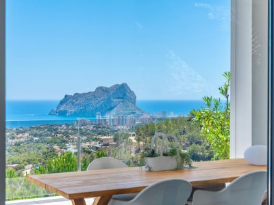 Buiten eetgedeelte met een houten tafel en moderne witte stoelen, met panoramisch uitzicht op de Peñon de Ifach, de Middellandse Zee en de kustlijn van Calpe.