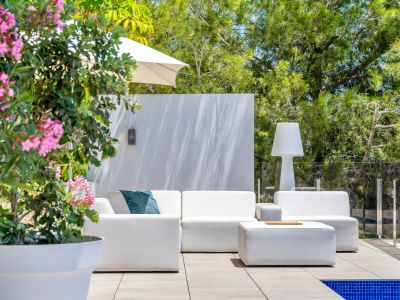 Hedendaagse buitenlounge met witte modulaire banken en bijpassende poef op een betegeld terras. Omgeven door weelderig groen, een glazen balustrade en een roze bloeiende plant.