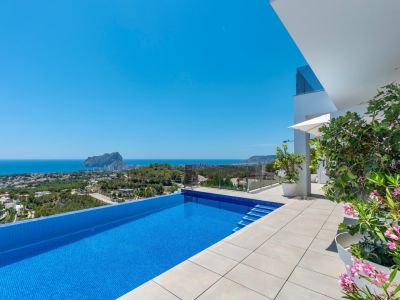 Infinity zwembad met blauwe mozaïektegels en ruim licht betegeld terras. Onbelemmerd panoramisch uitzicht op zee en de Peñon de Ifach, aangevuld met moderne architectuur en mediterrane flora.