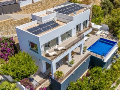 Luchtfoto van een moderne witte villa met meerdere terrasniveaus, een rechthoekig zwembad en ruime buitenlounges. Zonnepanelen zijn zichtbaar op de platte daken, omringd door weelderige mediterrane beplanting.