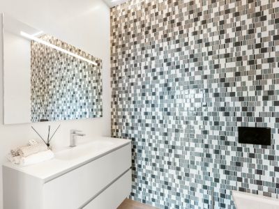 Moderne badkamer met een wit zwevend badmeubel en geïntegreerde wastafel. De accentmuur is bekleed met grijs-witte mozaïektegels, gecombineerd met een verlichte spiegel.