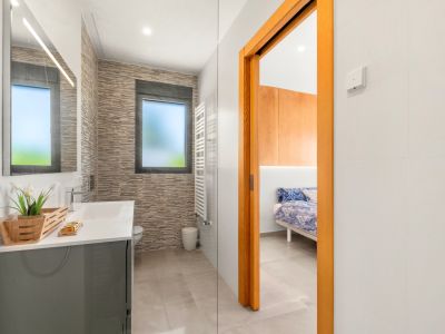 Moderne badkamer met grijze wastafel, verlichte spiegel en getextureerde wandtegels. Een matglazen raam en een witte handdoekradiator zijn zichtbaar, grenzend aan een slaapkamer.