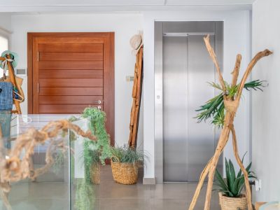 Moderne binnenhal met een strakke houten deur en een roestvrijstalen lift. De ruimte is elegant gedecoreerd met natuurlijke elementen zoals drijfhout, planten in manden en een glazen balustrade.