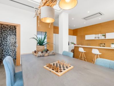 Moderne eetruimte met grijze tafel en lichtblauwe stoelen. Een unieke hanglamp van natuurlijk hout en een open keuken met lichte houten panelen zijn zichtbaar. Toegang tot een mozaïekte badkamer.