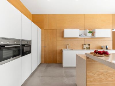 Moderne keuken met geïntegreerde witte kasten en Bosch-ovens. Het centrale eiland met licht stenen werkblad en de lichte houten wand- en plafondbekleding kenmerken de ruimte.