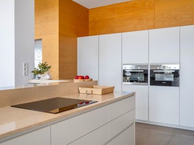 Moderne keuken met zandkleurig werkblad en geïntegreerde inductiekookplaat. Witte greeploze kasten herbergen twee ingebouwde Bosch-ovens en een houten accentmuur.