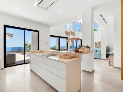 Moderne open keuken met centraal eiland, beige aanrechtblad en geïntegreerde inductiekookplaat. Grote glazen schuifdeuren bieden direct uitzicht op de Middellandse Zee, wat de lichte, eigentijdse ruimte versterkt.