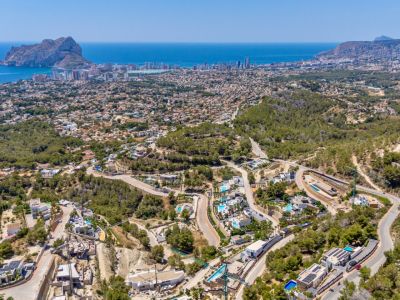 Panoramisch luchtbeeld van Calpe, met de Peñon de Ifach, de kustlijn en een residentiële wijk met villa's en zwembaden tussen groene heuvels en kronkelwegen.