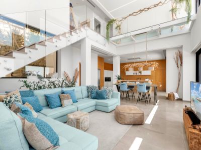 Ruime open woonkamer met lichtblauwe hoekbank, moderne glazen trap en keuken met warme houten accenten, rijkelijk verlicht door natuurlijk lichtinval.