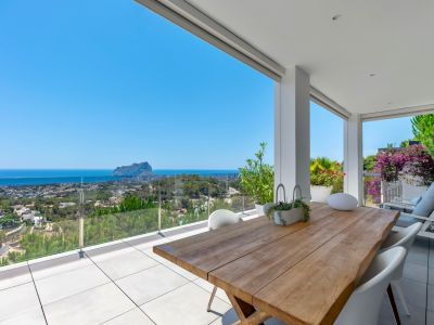 Ruime overdekte terras met een massief houten eettafel en witte stoelen. Biedt panoramisch uitzicht op de zee, Calpe en de Peñón de Ifach via een glazen balustrade.