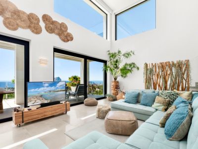 Ruime woonkamer met hoge plafonds, grote glazen schuifdeuren en bovenramen die panoramisch zeezicht bieden. Een lichtblauwe hoekbank combineert met natuurlijke houten accenten en wanddecoratie.