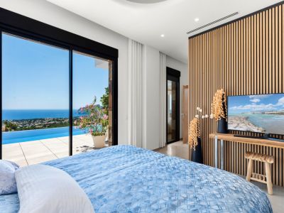 Slaapkamer met grote glazen schuifdeuren naar een infinity pool en panoramisch zeezicht. Een moderne houten lattenwand vormt de achtergrond voor de tv, passend bij het lichte interieur.