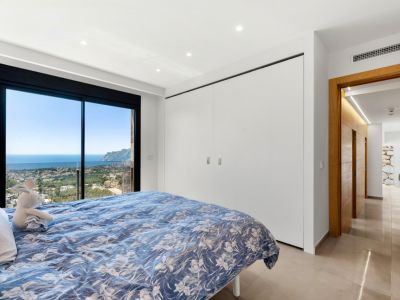 Slaapkamer met kamerhoge glazen schuifdeuren en panoramisch uitzicht op zee en stad. Voorzien van geïntegreerde witte kasten en een lichte tegelvloer.
