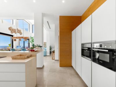 Strakke moderne keuken met geïntegreerde Bosch ovens, witte greeploze kasten en een licht stenen eiland. De open indeling verbindt met een eetruimte met panoramisch zeezicht via glazen deuren.