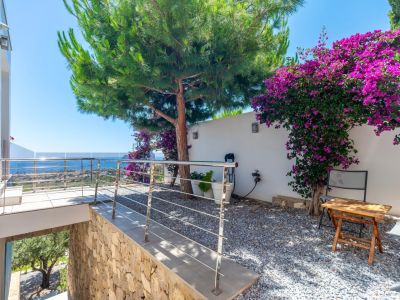 Terras met moderne RVS balustrade en glas, biedt panoramisch zeezicht. Aangrenzende grindtuin met grote dennenboom en levendige paarse bougainvillea tegen een witte muur.