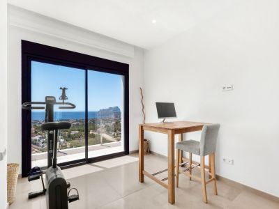 Veelzijdige kamer met houten hoog bureau en hometrainer, gericht op het weidse uitzicht op zee en de Peñon de Ifach door de grote glazen schuifdeuren.