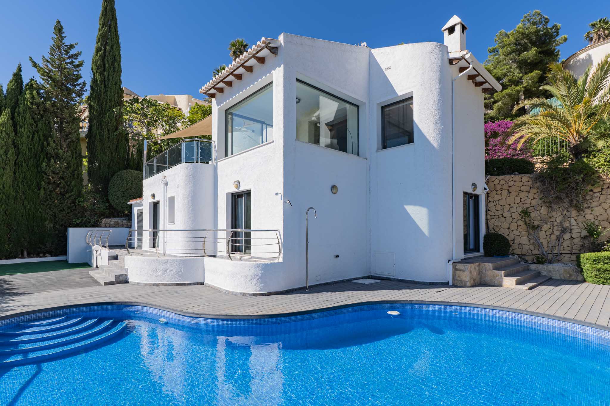 Buitenaanzicht van de villa, met een gebogen blauw mozaïekzwembad, grijze composiet vlonder, buitendouche en mediterrane beplanting.
