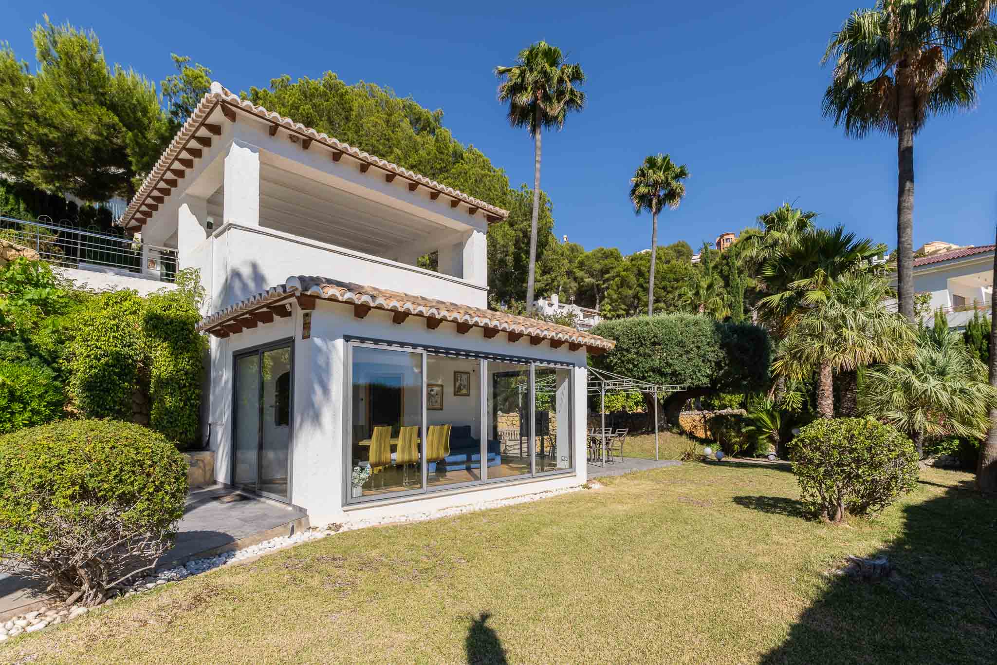 Buitenaanzicht van de villa: witte gevel, traditionele dakpannen, grote glazen puien. De vlakke tuin biedt een gazon en een terras met pergola, omgeven door palmen en struiken.