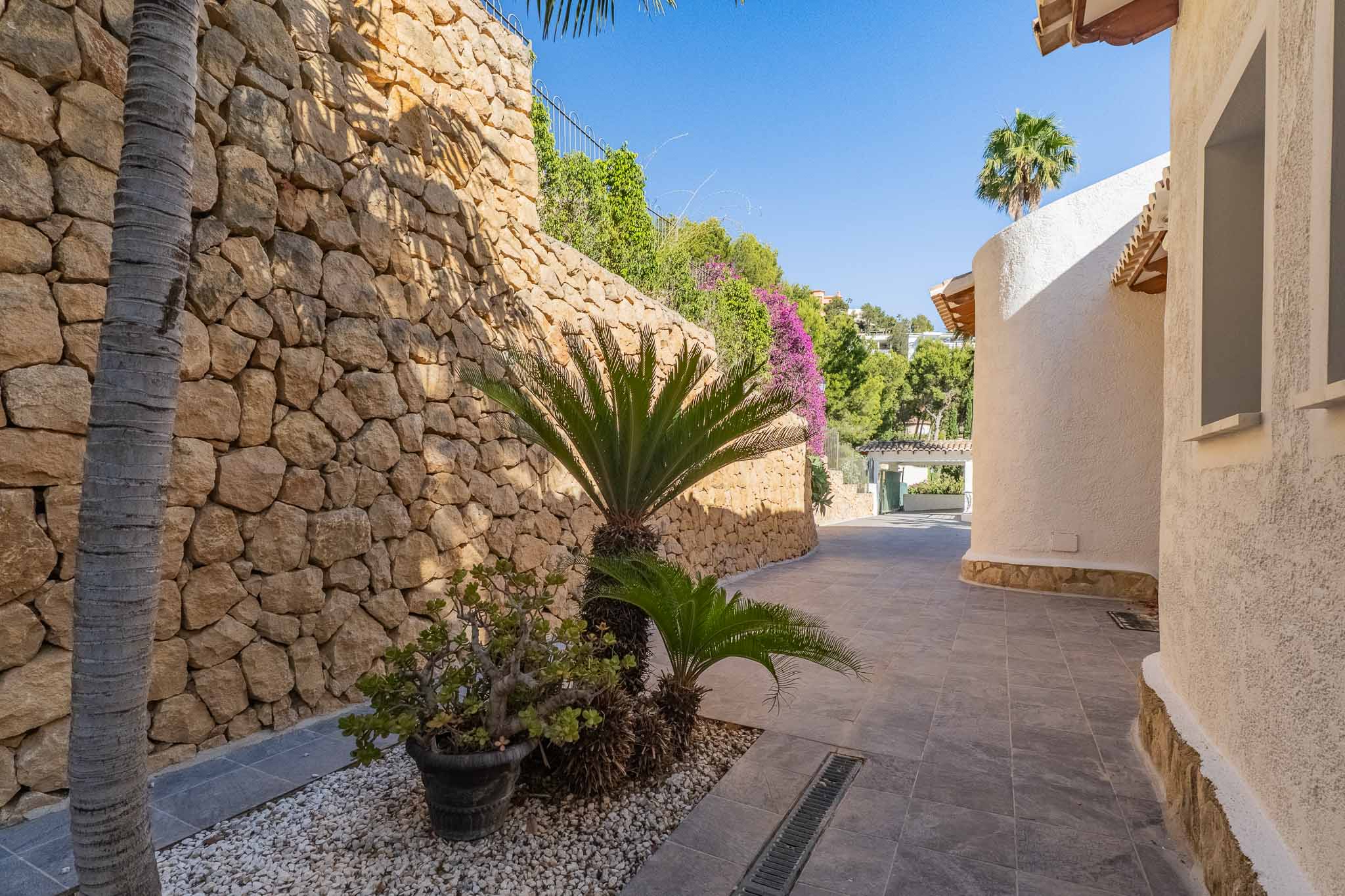 Buitenpad met robuuste natuurstenen keermuur en mediterrane beplanting, grenzend aan een gebogen witte stucgevel in fel zonlicht.
